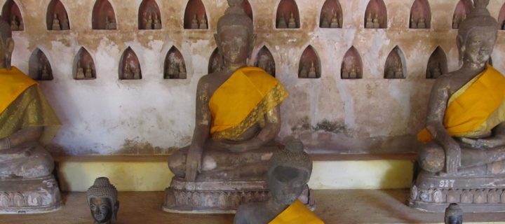 Photo shows six Buddha statues, three on a raised platform and three below. A scarf is draped over the left shoulder and wrapped under the right arm of each statue.
