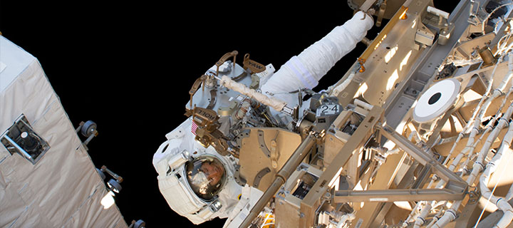An astronaut in a space suit appears to be tinckering with the outside of a satellite, both floating in outerspace.