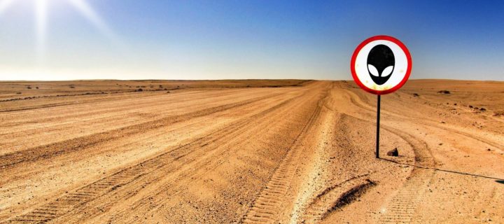 Tire tracks on a dirt road in the desert. To the right of the tracks is a circular signpost on a pole with the image of an extraterrestrials head.