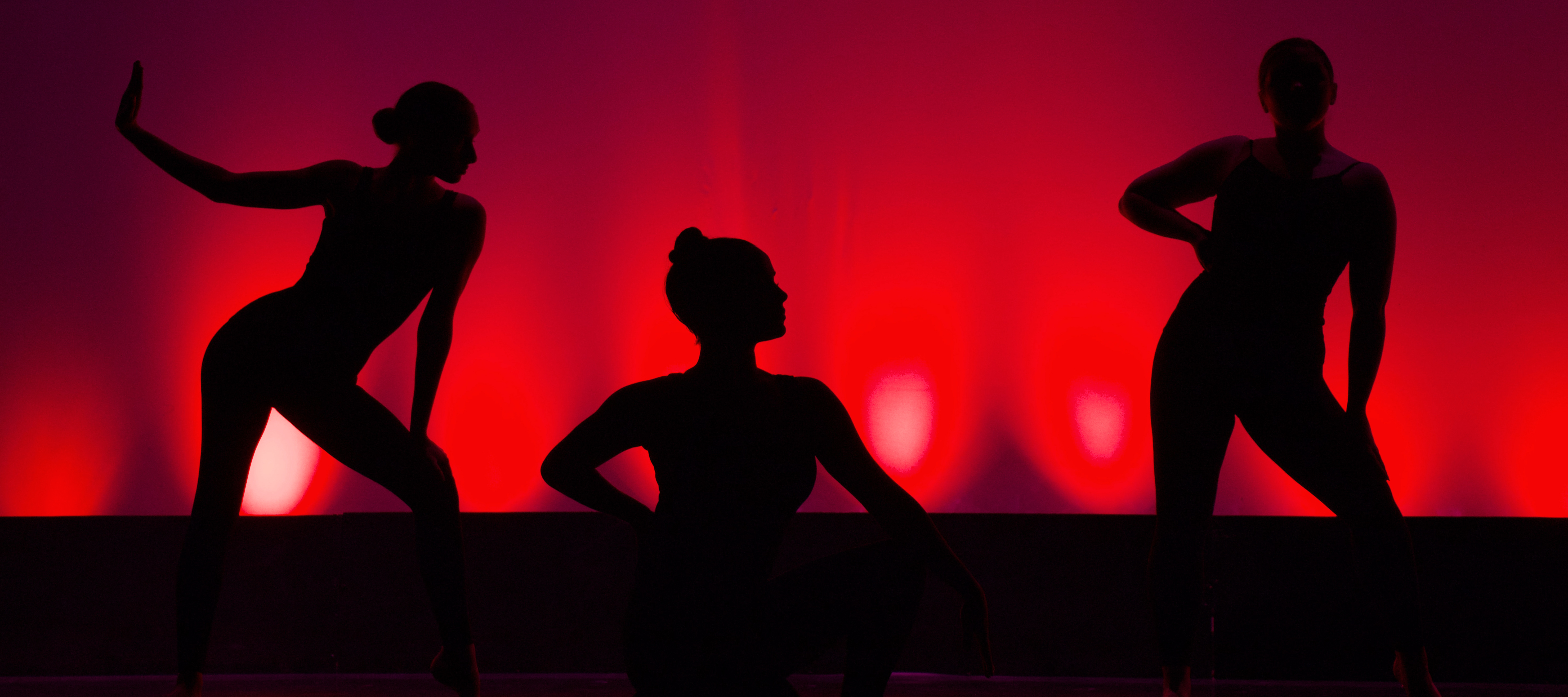 Three women dancers are silhouetted before a red background. The left-most dancer stands with one palm out away from herself and with the other on her knee. The middle danger kneels in profile. The right-most dancer stands with one hand on her hip and the other on her thigh.