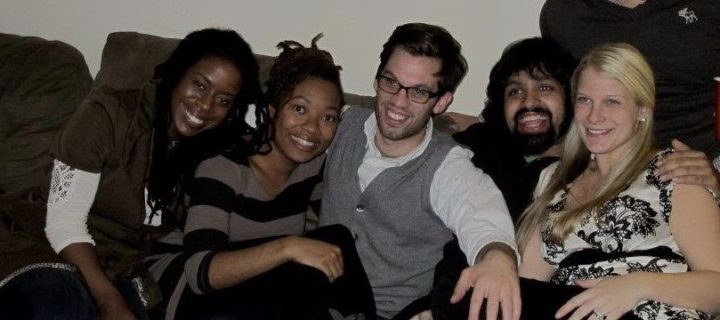 A photo of six people sitting on a couch leaning against each other and smiling.