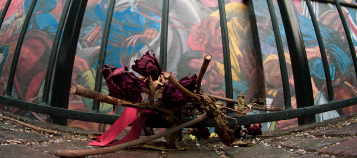 A photograph taken from the ground up showing a pile of dried flowers in front of the gate with part of the mural visible behind it.