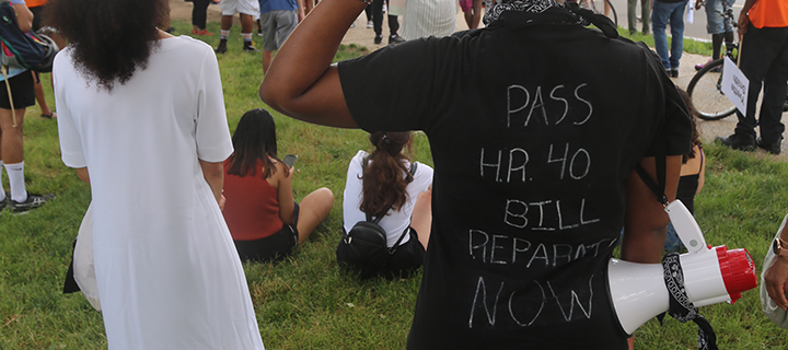 Photograph of people standing on grass, one person wearing a shirt that says "Pass H.R. 40 Bill Reparations Now."