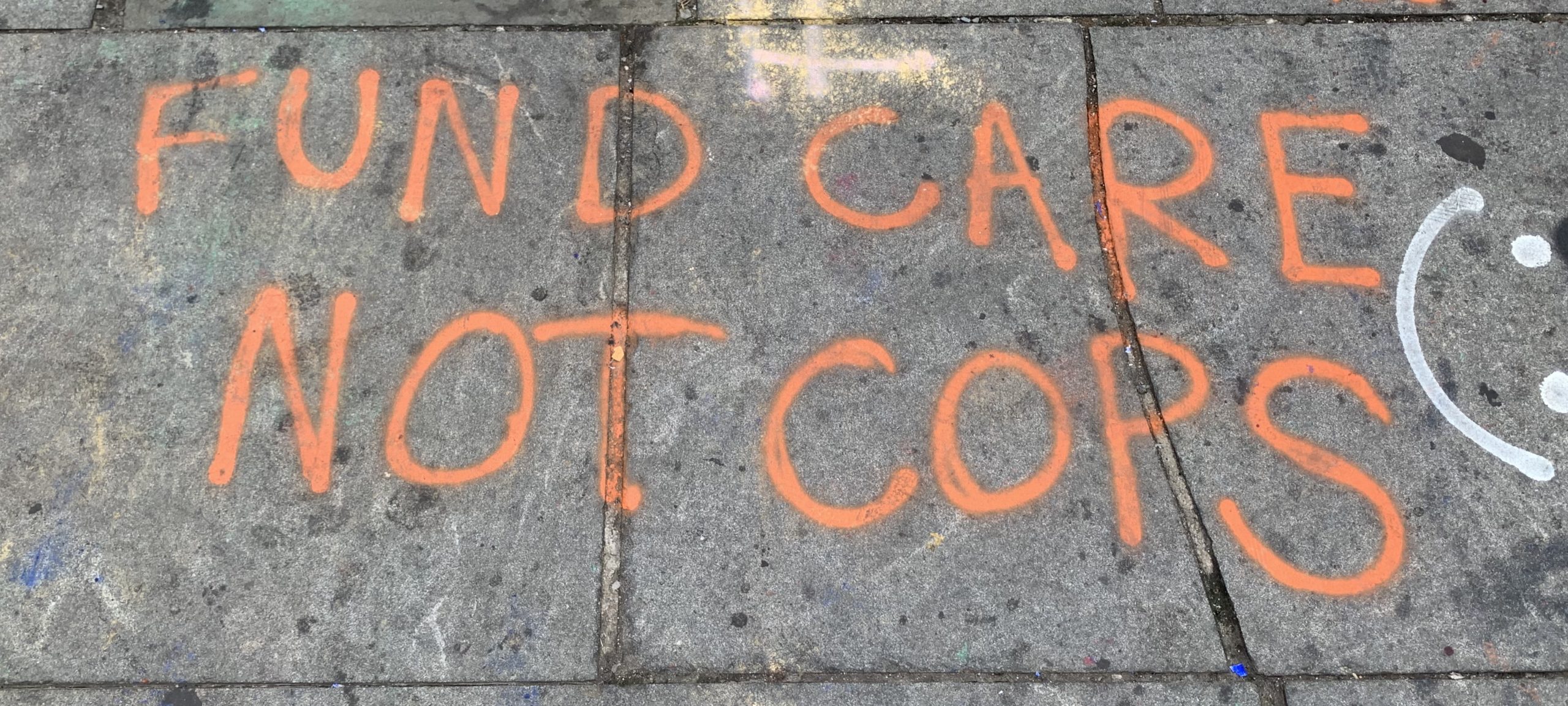 A picture of a message spray-painted onto a concrete sidewalk. The message says, "fund care not cops."